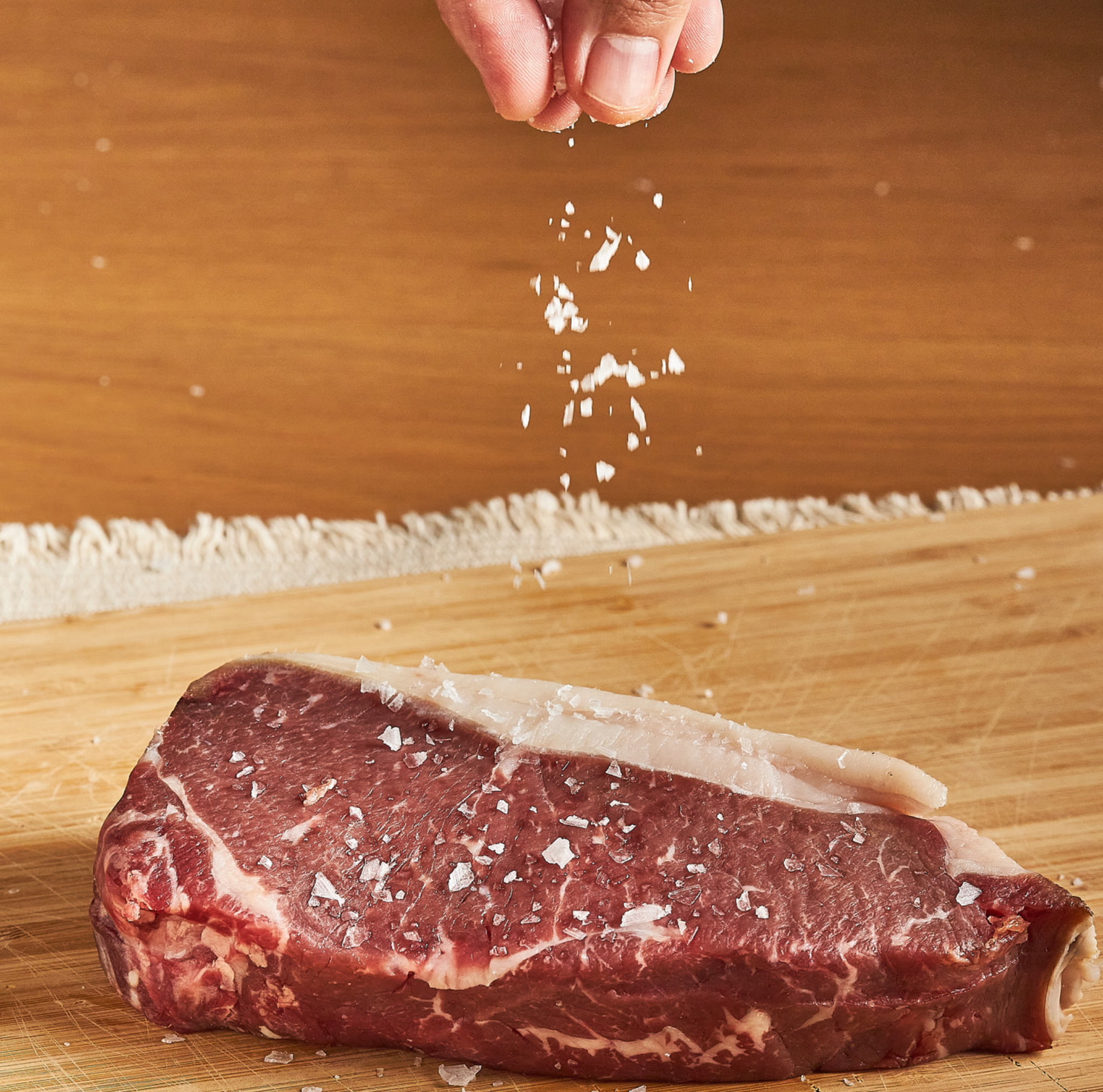 Raw steak on a wooden cutting board with salt being sprinkled over it.